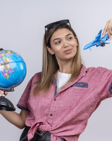 a pleased young woman wearing red shirt in sunglasses holding a globe while flying a blue toy plane on a white background